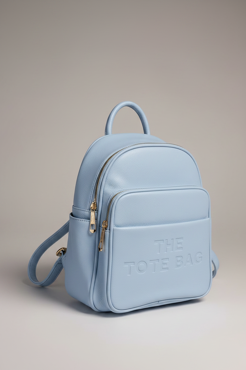 A light blue faux leather backpack with a top handle, side strap, and gold-tone hardware, featuring the embossed text 'THE TOTE BAG' on the front pocket, set against a neutral beige background.