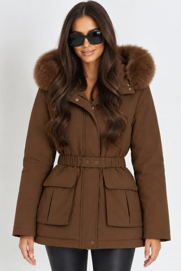 A woman wearing a brown hooded winter parka with a faux-fur trimmed hood, a matching drawstring waist belt, and large front flap pockets, photographed against a plain light gray studio background.