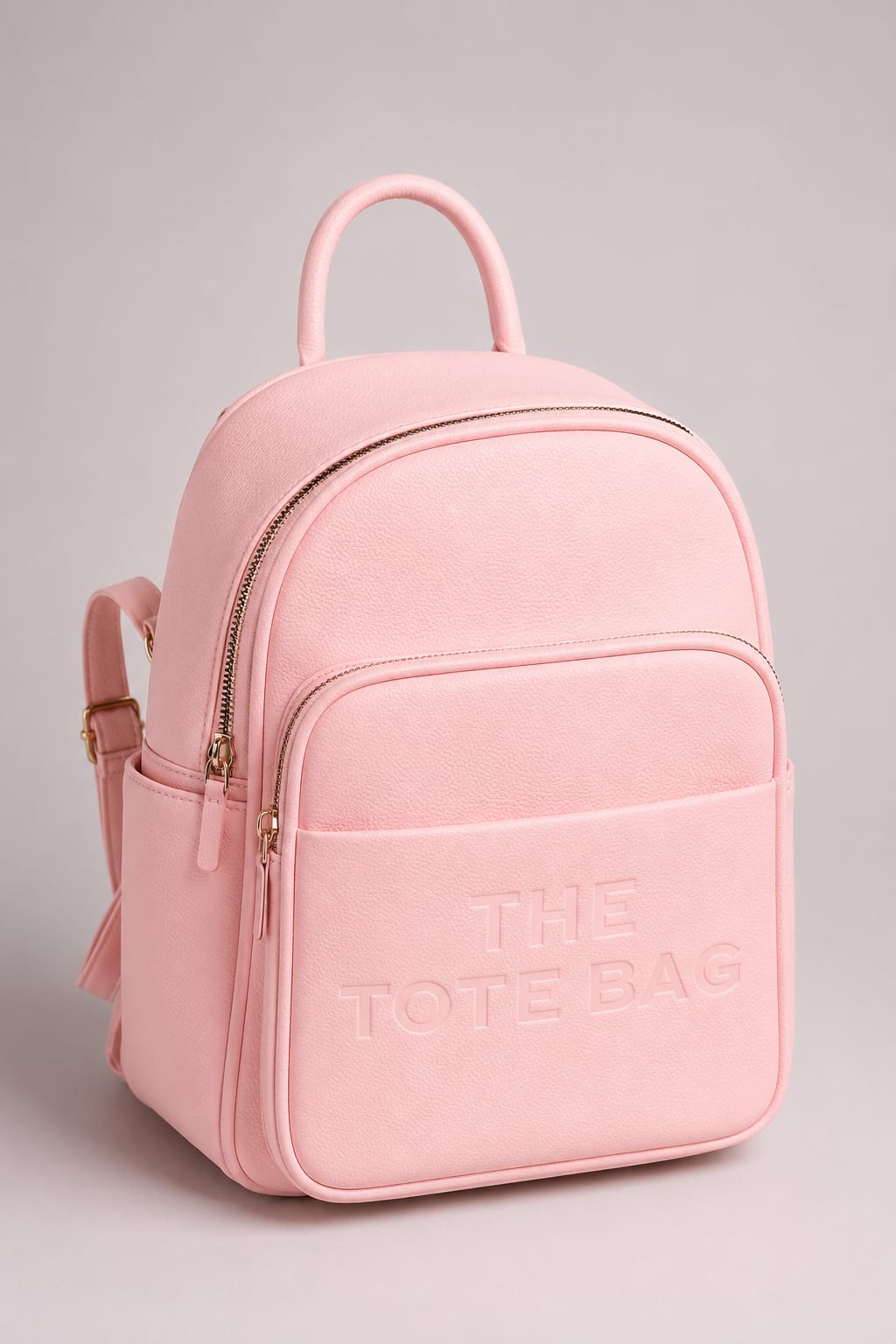 A light pink faux-leather backpack with a top handle, front zippered pocket, and side pocket, featuring the text 'THE TOTE BAG' embossed on the front, set against a neutral gray background.