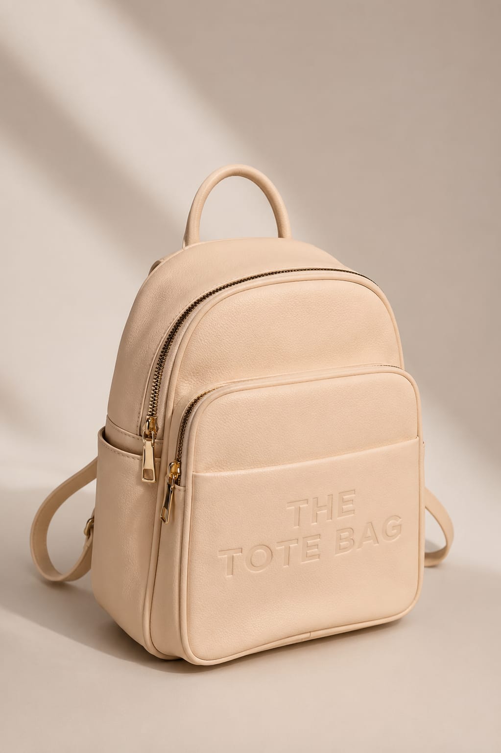 A light beige faux-leather mini backpack with gold-tone hardware, featuring an embossed logo that reads 'THE TOTE BAG' on the front pocket, set against a warm beige background with soft diagonal shadows.