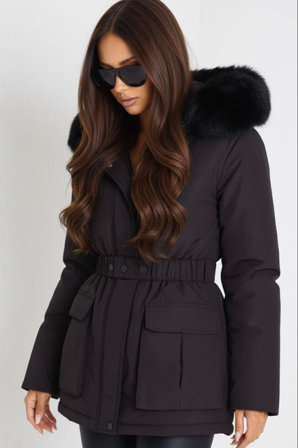 A woman wearing a black belted puffer parka with a black faux-fur hood, styled with black sunglasses against a light gray studio background.